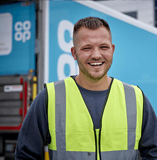 An employee in a high-visibility vest stands near a Co-op branded truck in a commercial area, with other vehicles and buildings in the background.