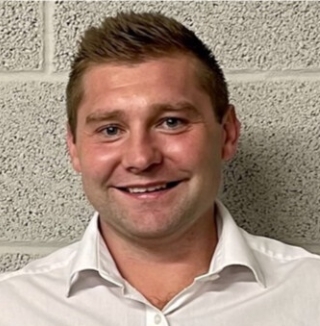 Man in white shirt smiling in front of brick wall.