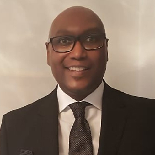 Man in black suit smiling in front of white wall.