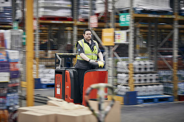 Person wearing a high-visibility vest and gloves operating a red and black electric pallet truck in a warehouse with shelves stocked with goods and section signs in the background