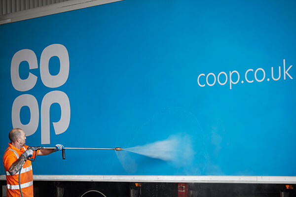 Person in an orange safety vest pressure washing a blue Co-op truck with visible branding.