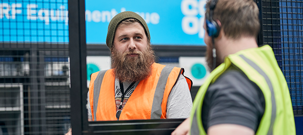 Two warehouse workers in high-visibility vests, separated by a metal mesh barrier, with blue signage in the background.