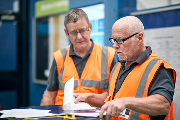 Two people in orange safety vests at a table with papers and tools, in front of a wall chart.