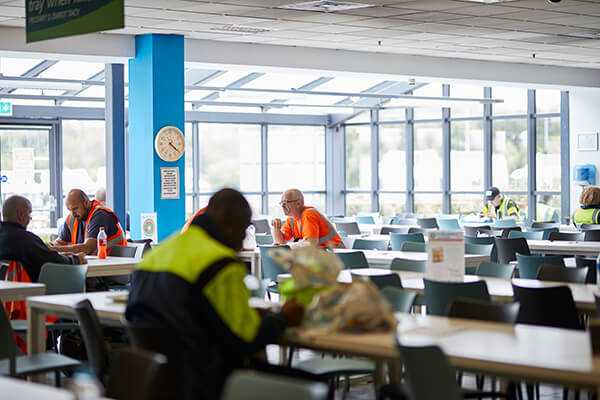 Workers in high-visibility clothing seated at tables in a bright cafeteria with a blue pillar and clock.