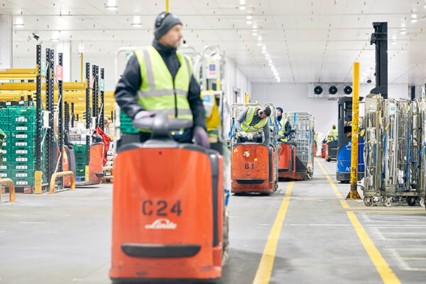 Workers in high-visibility vests operating orange pallet trucks in a well-lit warehouse.