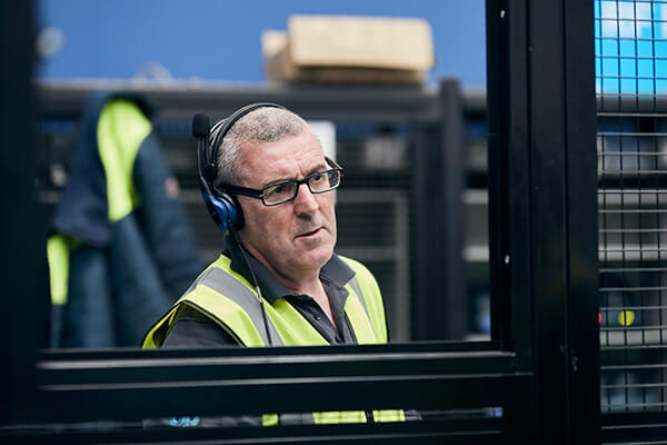 Person in a high-visibility vest and headphones behind a black metal barrier in a warehouse