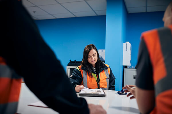 Three people in orange safety vests seated at a table with documents in a blue-walled room