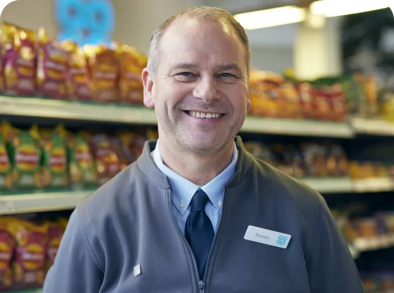 Individual in a grey jacket with a name tag, standing in a grocery store aisle with shelves of packaged goods in the background