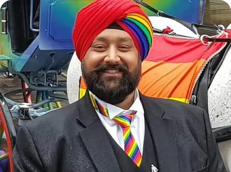 Individual in a black suit with a rainbow tie and turban, standing in front of a colorful background
