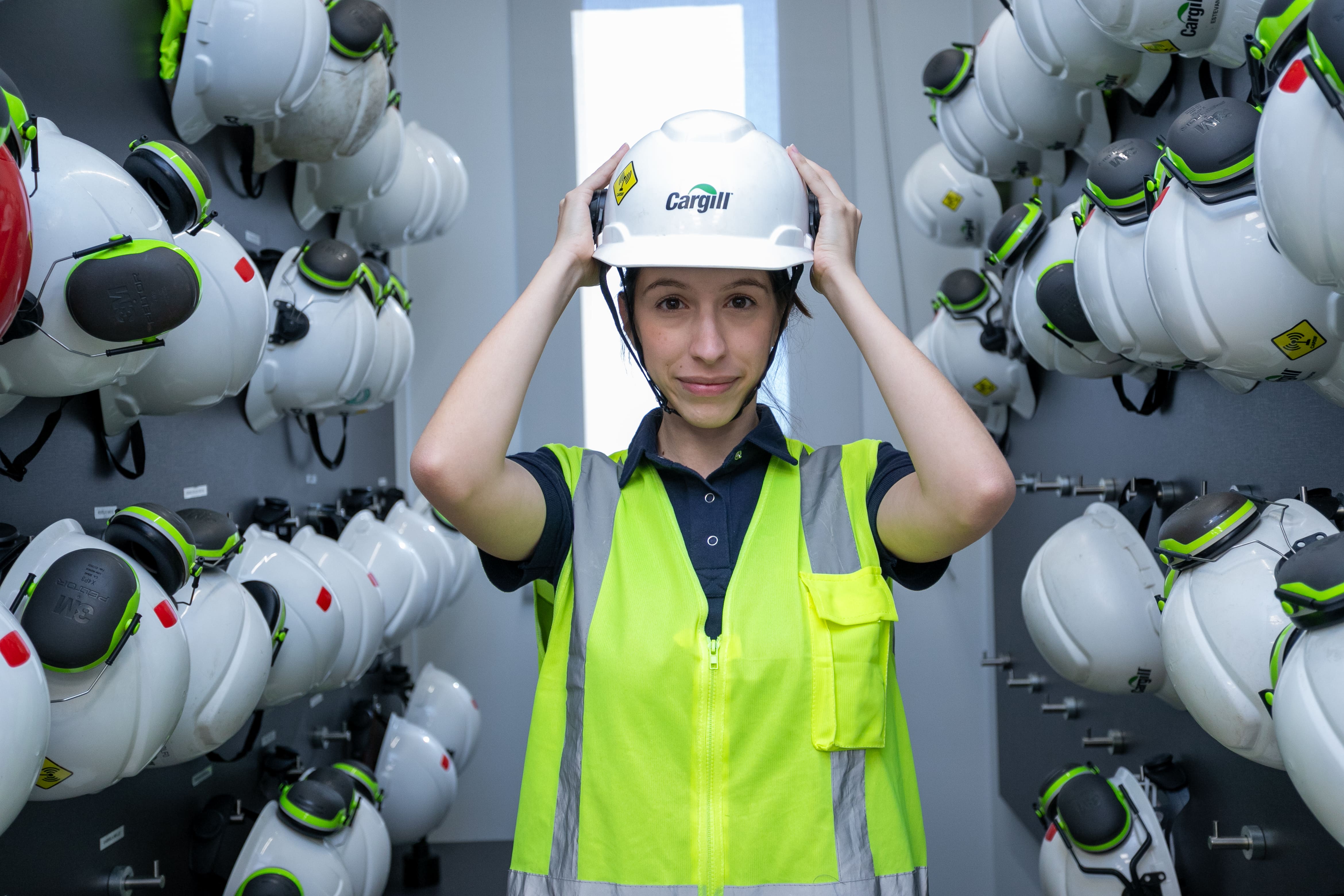 Colaborador de Cargill ajustando un casco de seguridad y usando chaleco de alta visibilidad en una instalación industrial, con equipo de protección personal almacenado al fondo.