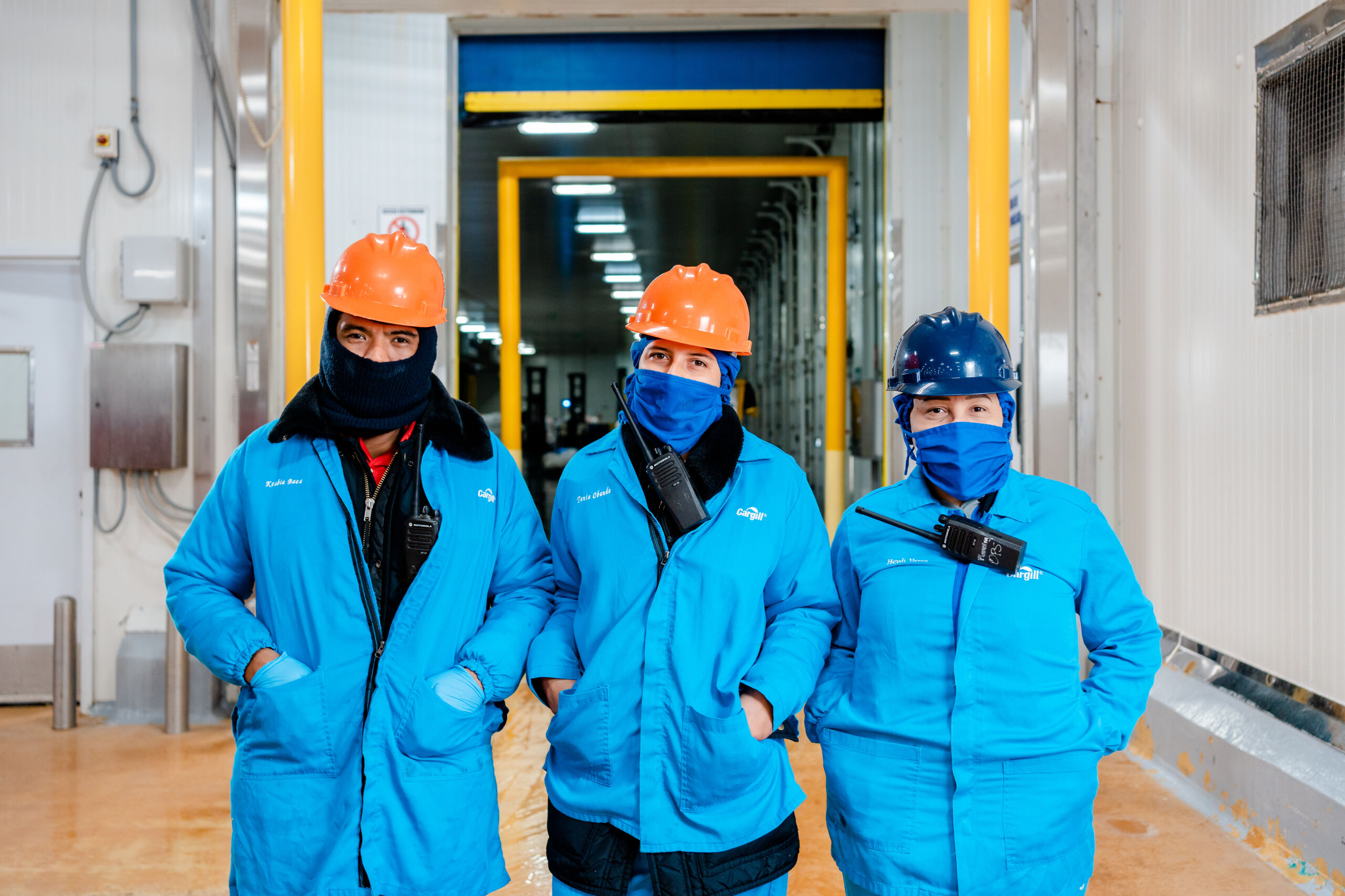 Tres colaboradores de producción de Cargill con cascos, ropa de protección y cubrebocas en una planta de manufactura, destacando la seguridad laboral y el trabajo en equipo.