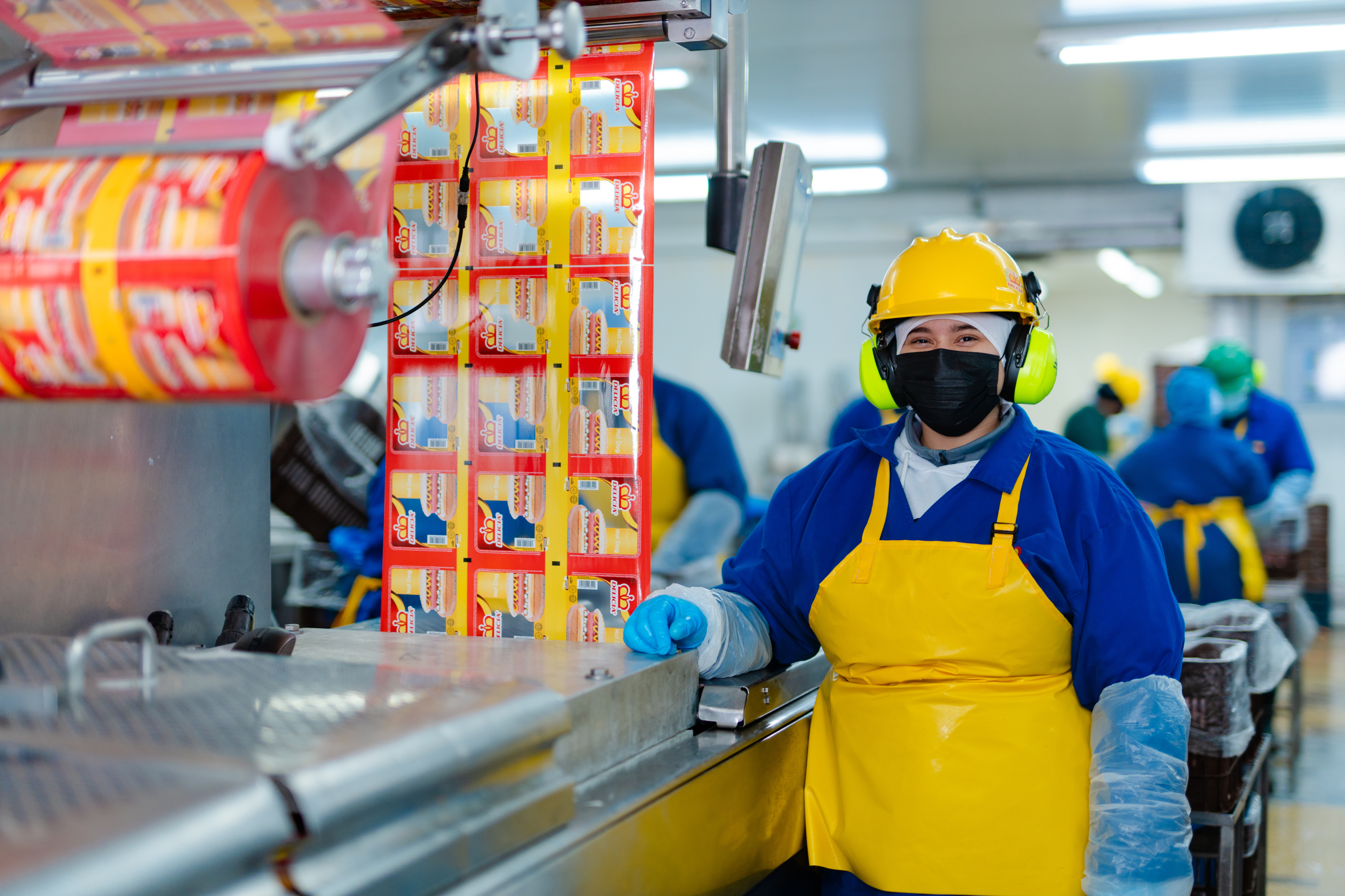 Colaborador de Cargill con casco, protección auditiva, mascarilla y ropa de protección junto a equipos de empaque en una planta de producción de alimentos.