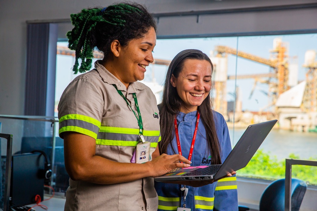 Two Cargill employees reviewing information on a laptop while collaborating inside an industrial facility with production equipment visible in the background.
