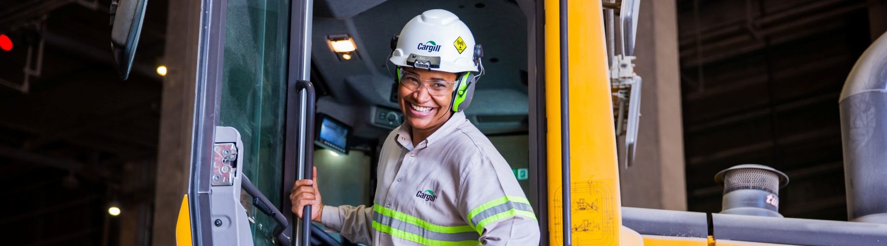 Cargill employee wearing safety gear standing on the steps of a large yellow industrial vehicle inside a production facility.
