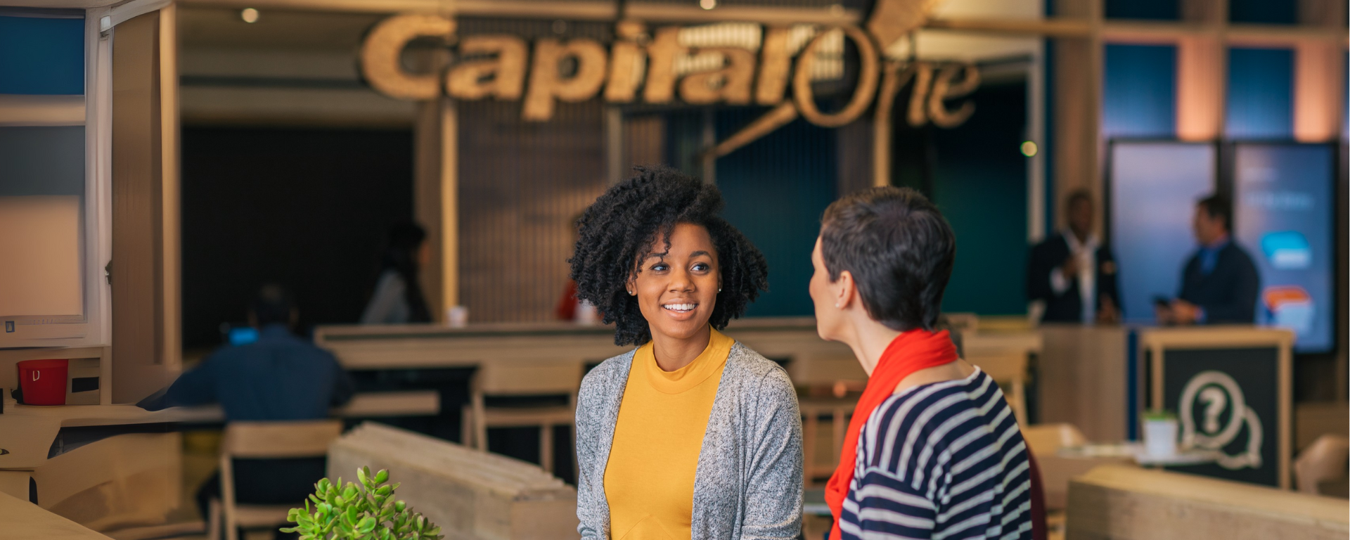 Two women wearing bring clothing and sweaters chat over coffee at a Capital One Café