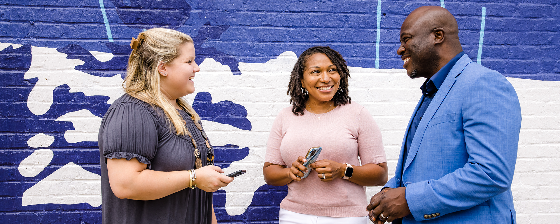 Three Capital One associates smile and talk in front of a blue and white brick wall