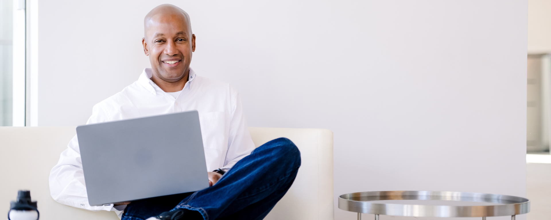Capital One associate sitting on a white sofa chair with ankle crossed over knee and holding a laptop in lap
