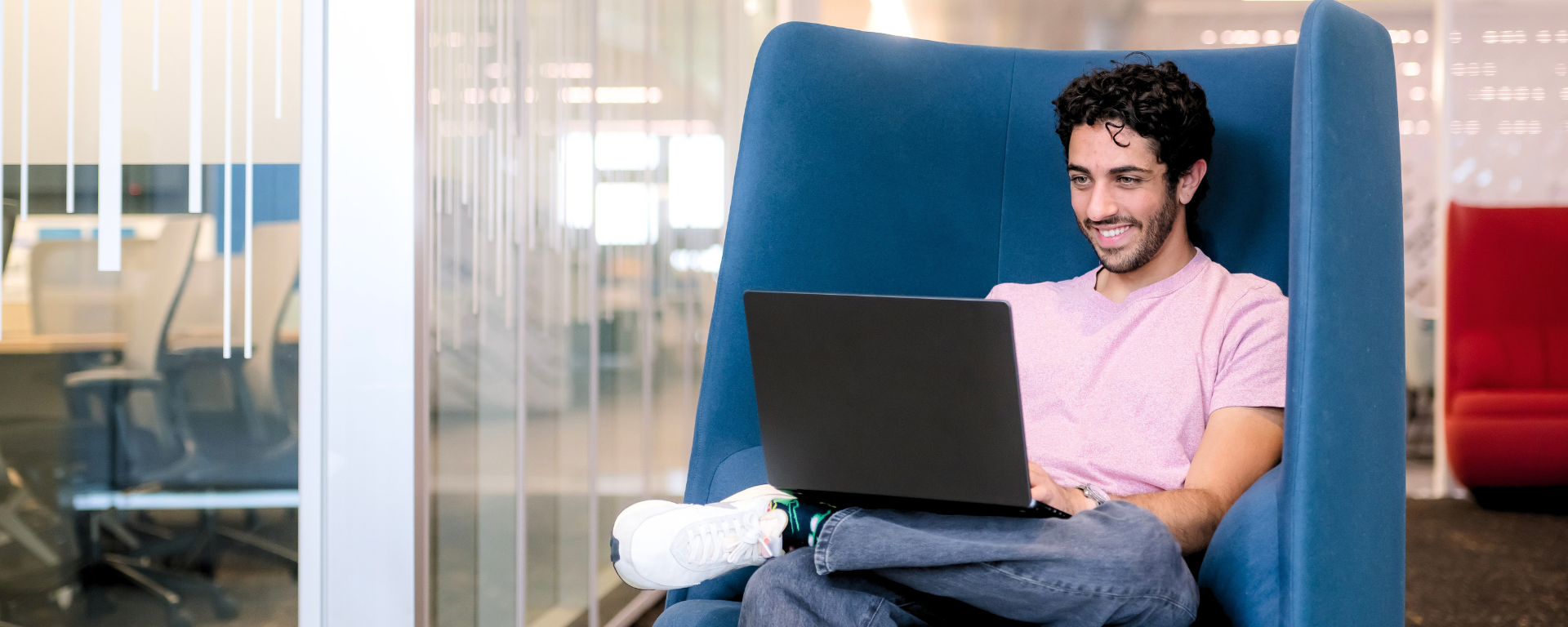 Capital One associate sitting in a blue sofa chair wearing a pink shirt and jeans while working on a laptop in his lap