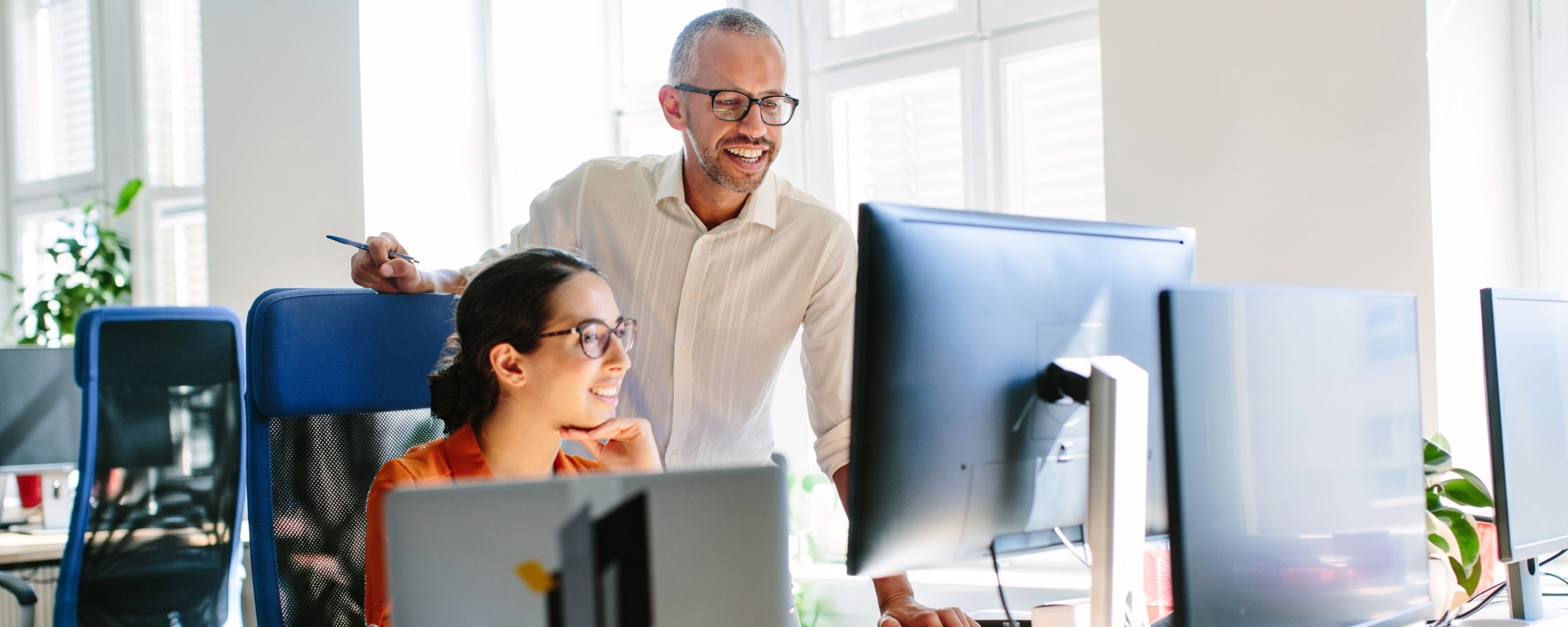 An associate wearing a white button down shirt and glasses looks over a peer's shoulder at her monitor. She is wearing an orange blouse and wears glasses.