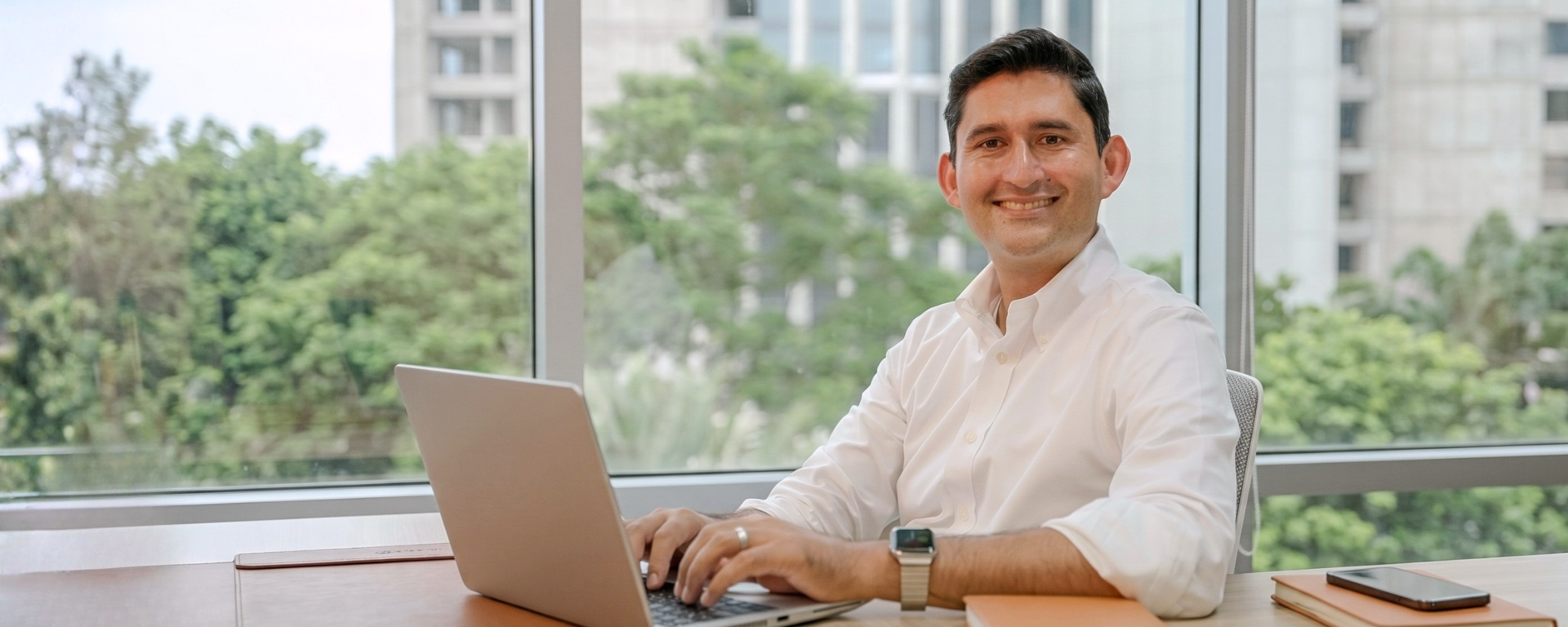 Capital One associate Oz sits at a desk by the window and types on his laptop while smiling at the camera