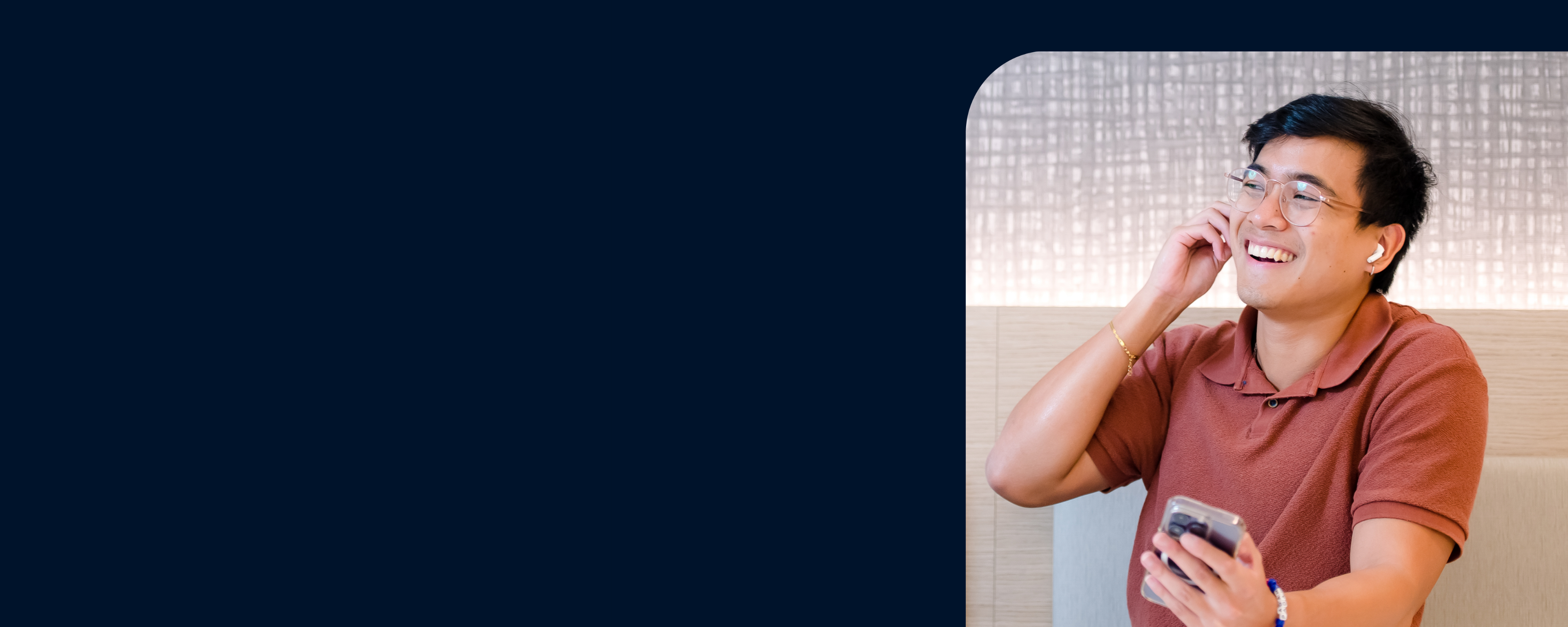 Photo of a Capital One associate sitting at a table booth laughing while holding a phone and wearing earbuds over a blue backdrop
