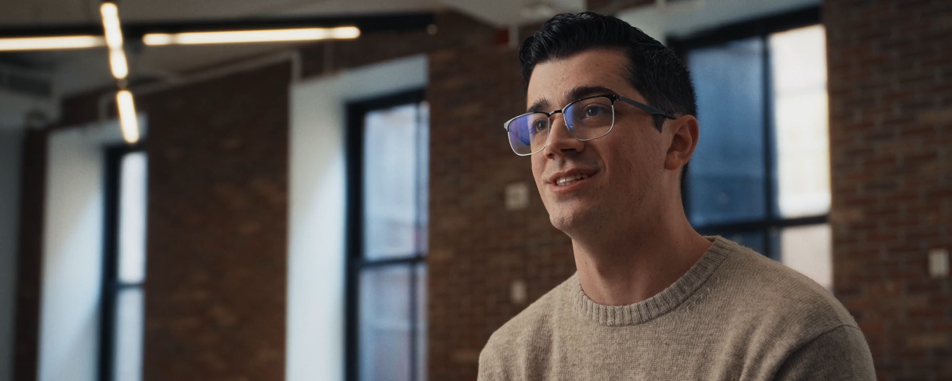 Capital One Students & Grads associate Lorenzo sits and talks in a room with brick walls wearing a tan long sleeve shirt and glasses