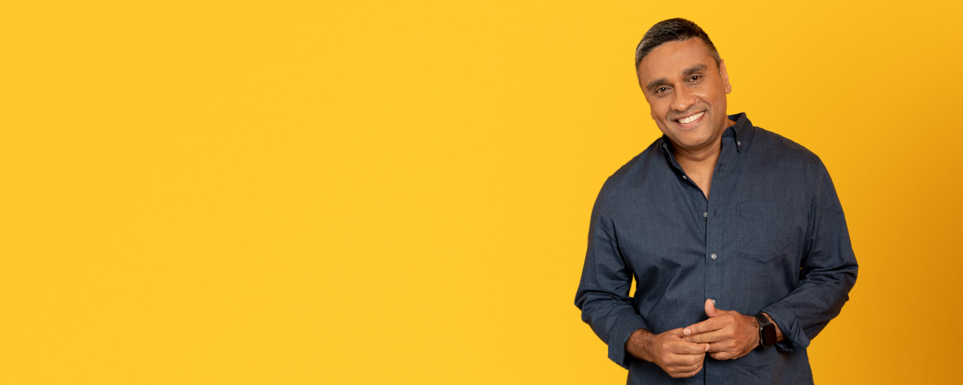 Capital One associate Salim stands and smiles in front of a yellow wall wearing a dark buttoned down shirt