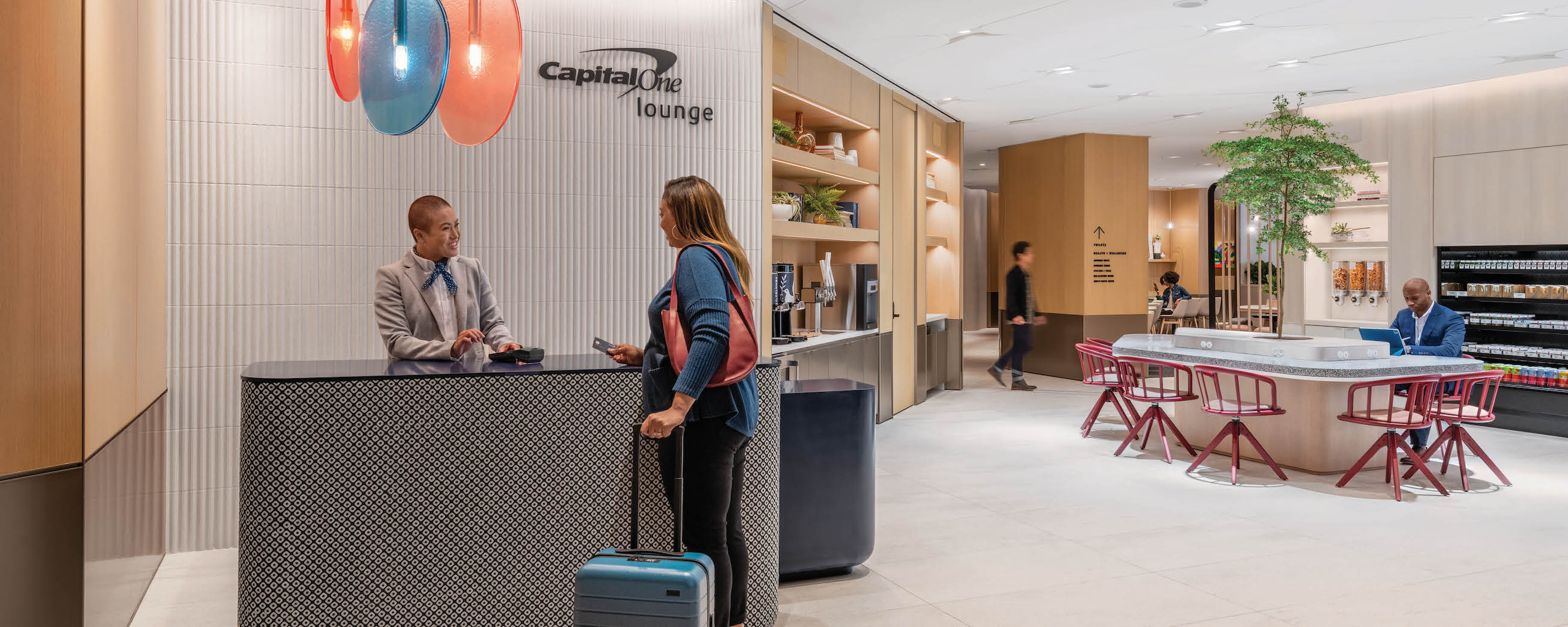 Woman with blue vest and suitcase is greeted by Capital One Associate at the Capital One airport lounge