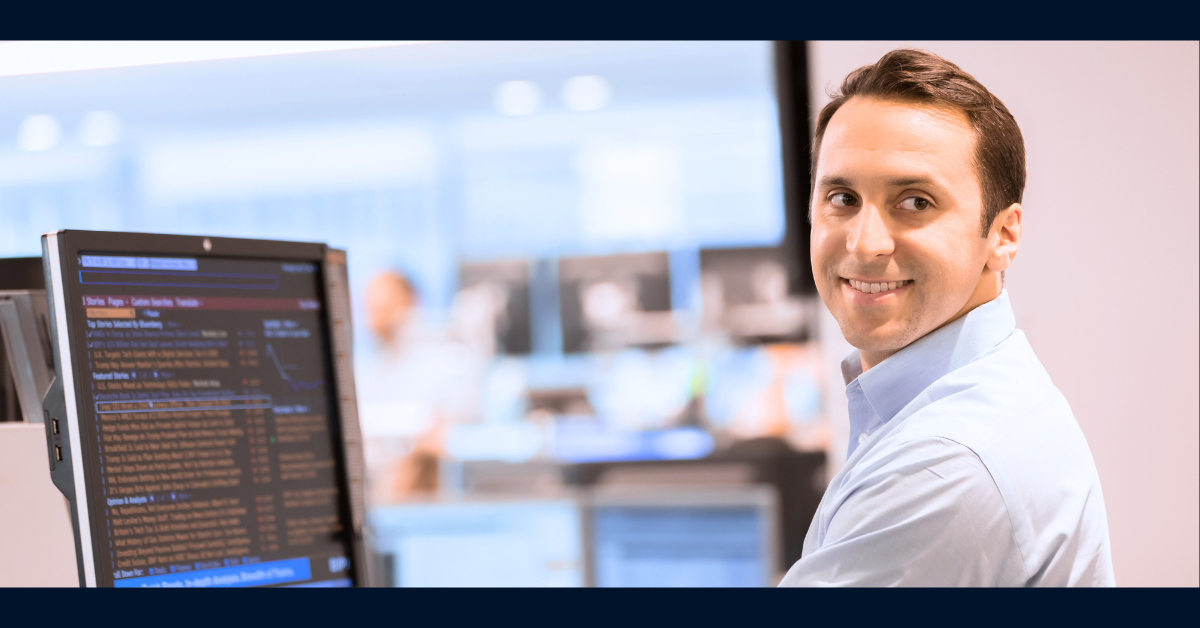 Capital One associate wearing a button up shirt turns to look over his shoulder and smiles as he is working on his monitor