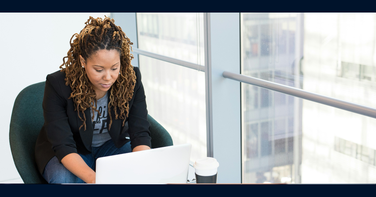 Capital One associate wearing a blazer sits in a green sofa chair and works on her laptop with a cup of coffee