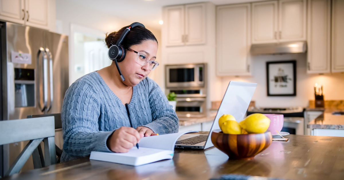 Capital One Customer Care associate works from her kitchen table, looking at her laptop and writing in a notebook