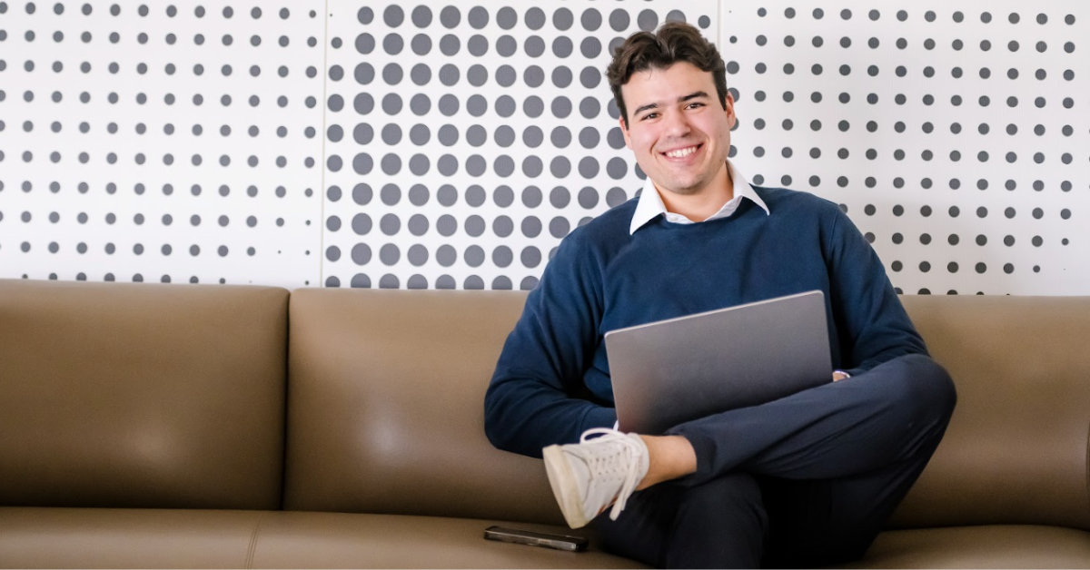 A Capital One associate sits on a brown couch holding his laptop in his lap and smiles