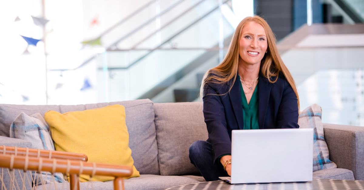 A Capital One associate sits on a gray couch and works on her laptop smiling