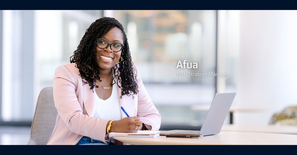 Afua, Tech Strategy Manager at Capital One, wears a white shirt and pink blazer while smiling and writing on a notepad with a blue pen