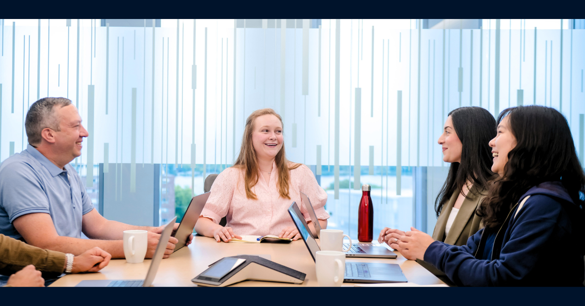 Four Capital One software engineers sit around a square table in a meeting room and laugh amongst each other while working on their laptops