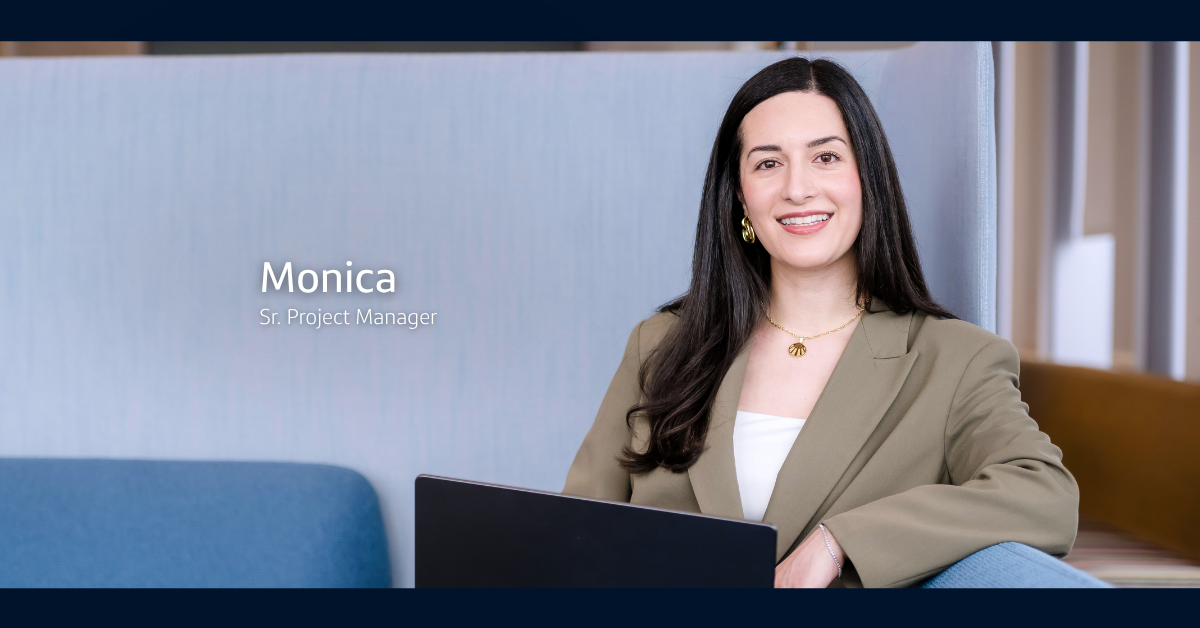 Monica, Sr. Project Manager at Capital One, smiles at the camera as she wears a brown blazer and leans against a blue couch