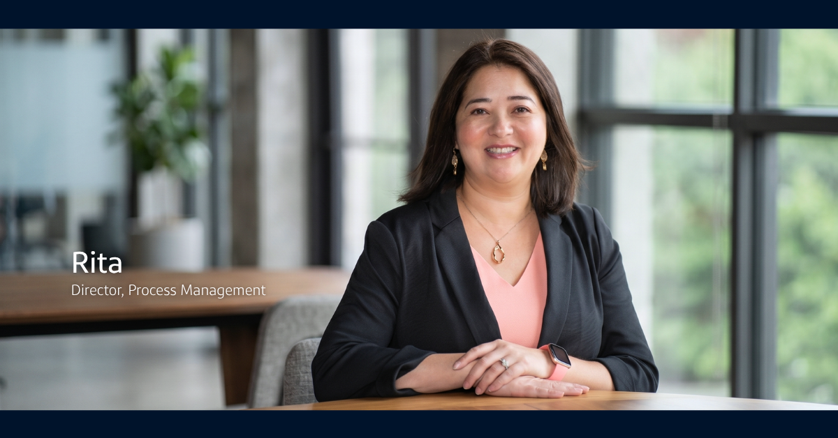 Rita, Process Management Director at Capital One, smiles at the camera while wearing a gray cardigan and pink blouse with her hands folded atop a wooden desk