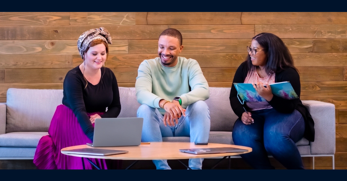 Three Capital One associates sit together on a gray sofa, smiling and looking at each other's work