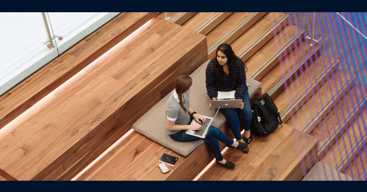 Two Capital One associates sitting on a wooden stairway with their laptops and talking
