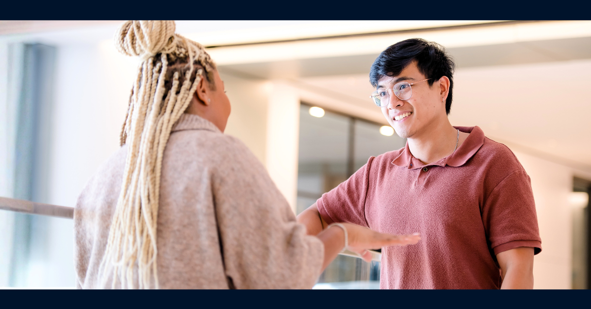 Two Capital One associates talk to one another in the office. Facing the camera is an associate wearing a red polo shirt and glasses, while the associate facing away from the camera wears a beige cardigan and blond braids