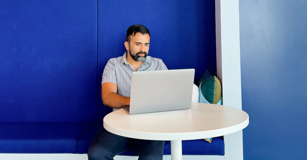 Capital One Product Manager Samir sits at his laptop in front of a royal blue wall inside a Capital One office