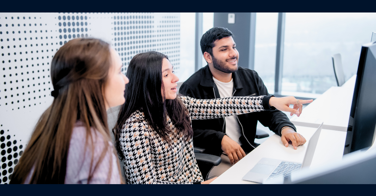 Three Capital One associates sit at an open desk, with the associate in the middle smiling and pointing to a monitor