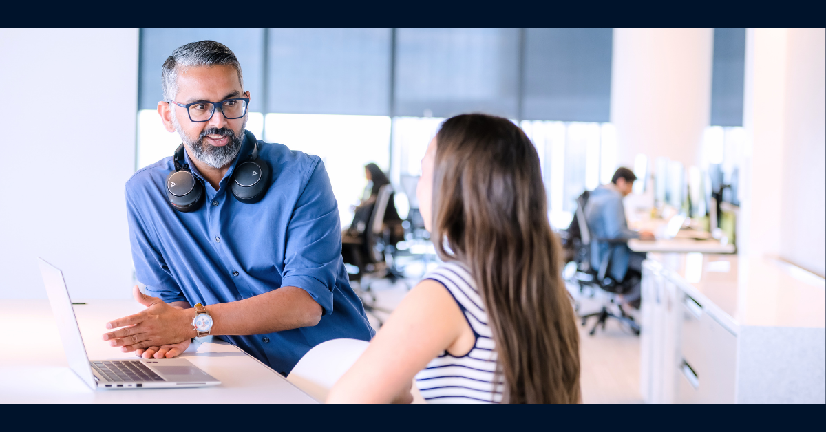 Capital One associate wearing a blue button down shirt gestures towards his laptop while speaking to a peer
