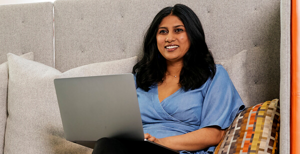 A smiling woman works on a laptop while seated on a couch with pillows