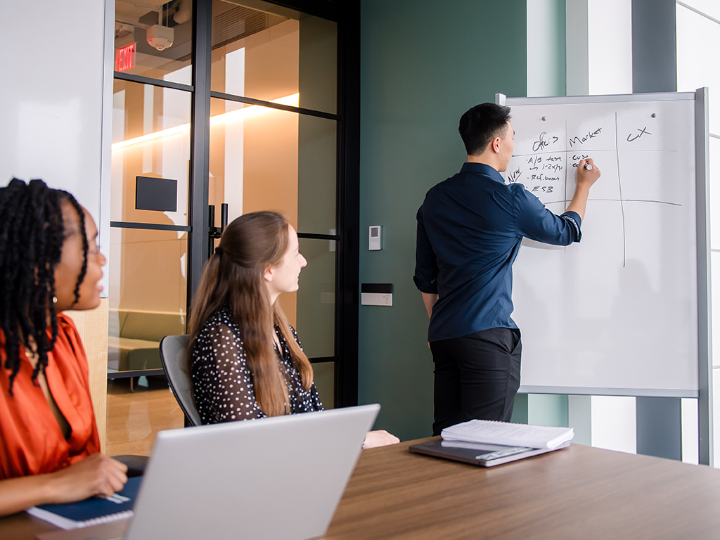 Three Capital One Strategy Consulting associates work in a room together, two sitting at a table and one writing on a whiteboard