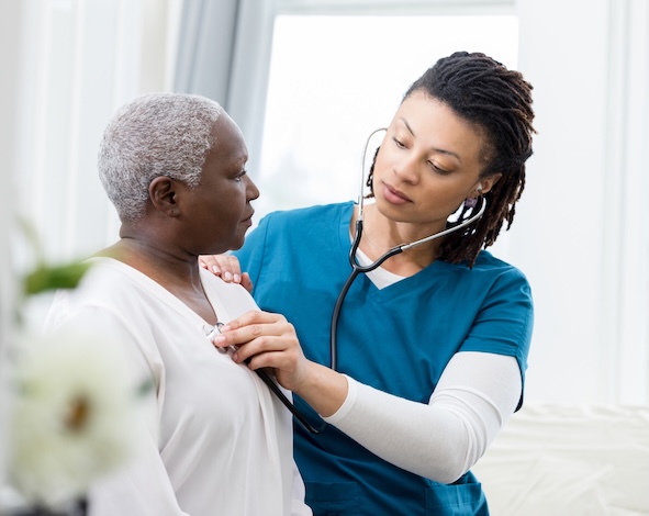 A caring mid adult female home healthcare nurse uses a stethoscope to check the heart and lungs on a senior female patient.
