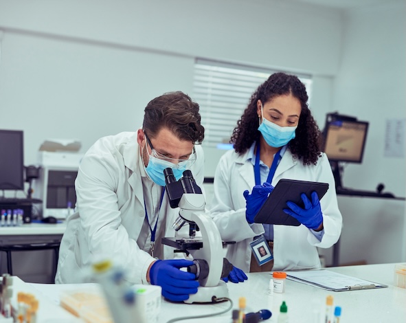 Female scientist and male colleague wearing facemask and gloves, focusing on work in laboratory, using digital tablet and microscope