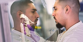 A person with a beard and short hair, wearing a lab coat, looks intently at something inside a clear enclosure, with their reflection visible on the surface.