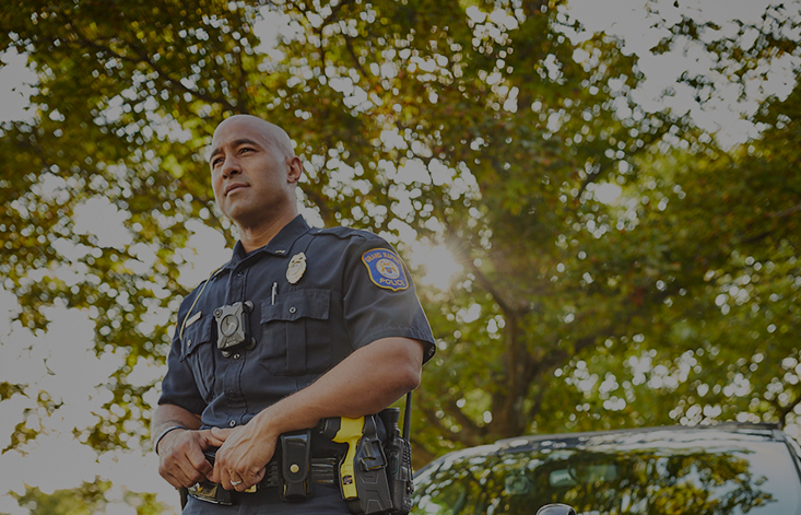 A police officer stands in front of a tree with both hands on his belt