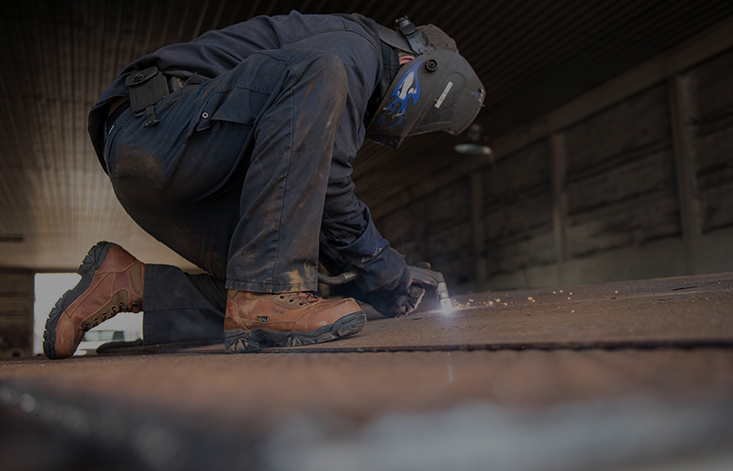 A welder with his visor down crouches to repair a piece of metal, sparks flying